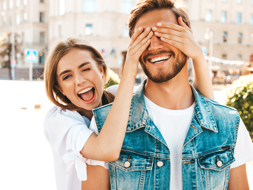 Smiling beautiful girl and her handsome hipster boyfriend. Woman covering her man eyes with hands. Happy cheerful family. Hugging loving couple standing outdoors.Guess who concept