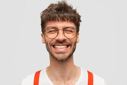 Positive bearded male hipster smiles broadly, has pleased expression, laughs at something funny, closes eyes, has trendy hairdo, isolated on white wall. Cheerful successful young businessman
