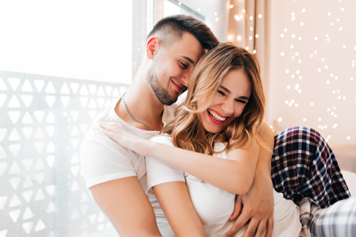 Excited girl with wavy hair having fun while boyfriend embracing her. Indoor photo of inspired guy expressing love.