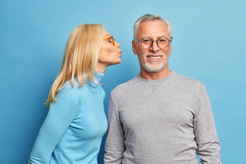 Affectionate elderly blonde woman going to kiss bearded husband in cheek expresses love. Two happy married people in casual wear have good relationship pose together against blue background.